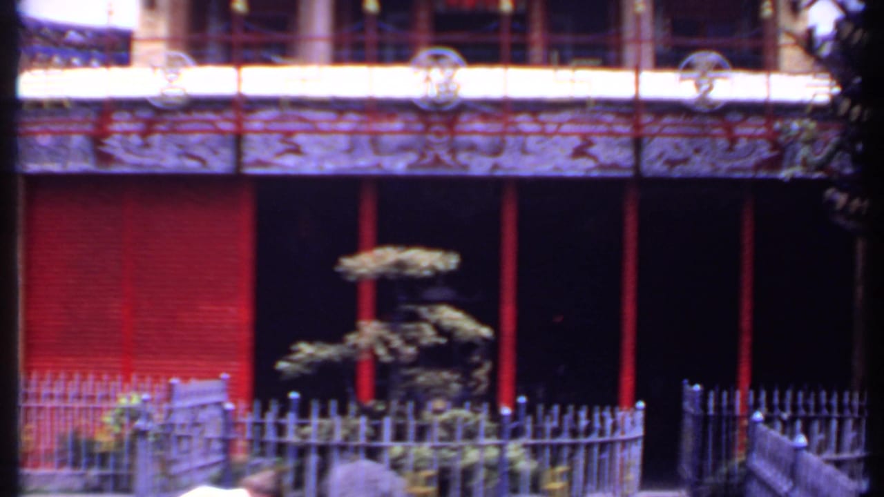 Handheld 1969 India-listed frame showing the temple's red lower facade, dark entry bay, courtyard fence, small shaped tree, and blurred visitors at the bottom.