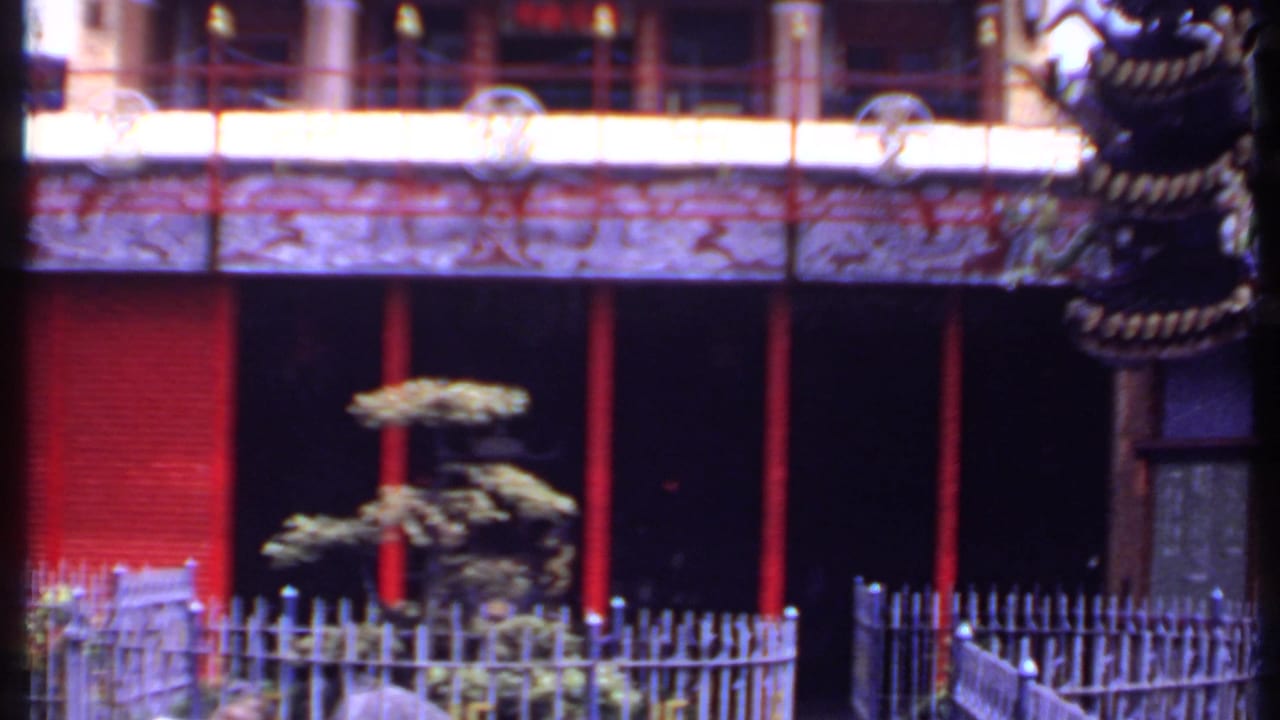 Temple facade with red pillars, carved trim, railings, and a small shaped tree in the courtyard.