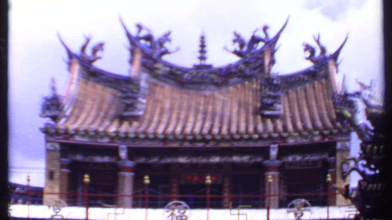Ornate temple roof with upturned eaves, sculptural ornaments, red railing, and cloudy sky.