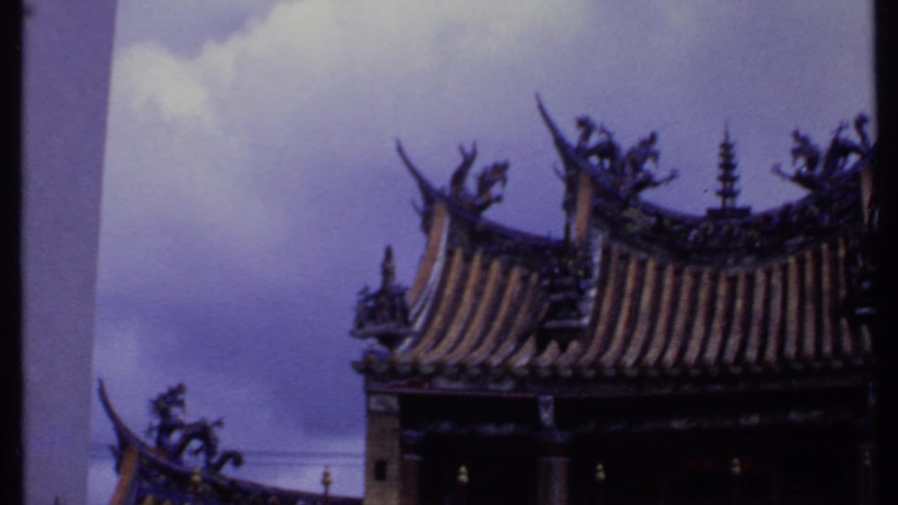 1969 India-labeled frame from the pan showing a partial temple roofline with upturned eaves, ornamental figures, dark facade, and open sky.