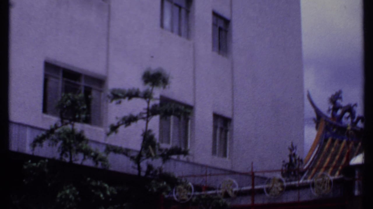 1969 India 8mm frame of a white urban building with trees, fencing, and an ornate roof edge at the side.