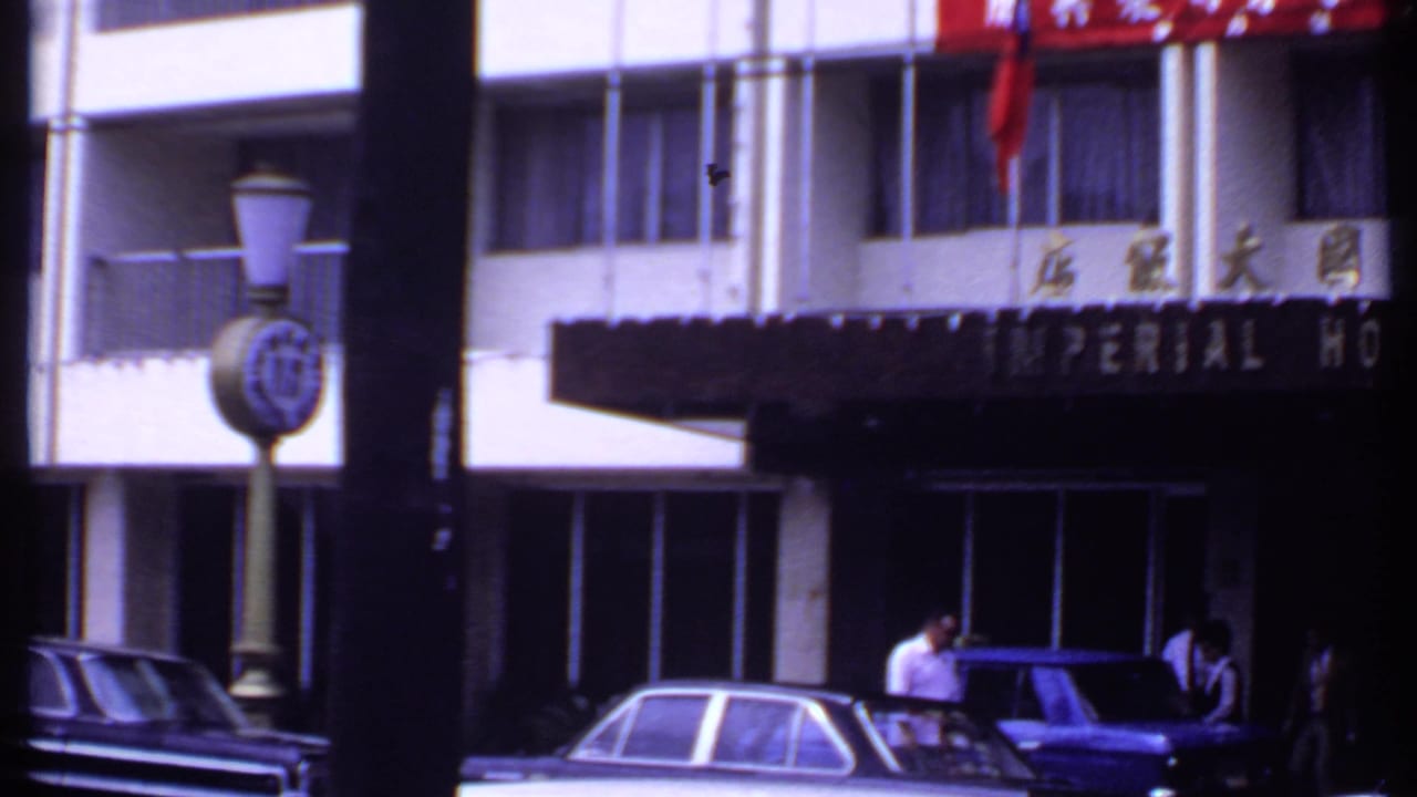 1969 India 8mm view of the Imperial Hotel exterior with a clock post in front, Chinese characters above, and people near parked cars.