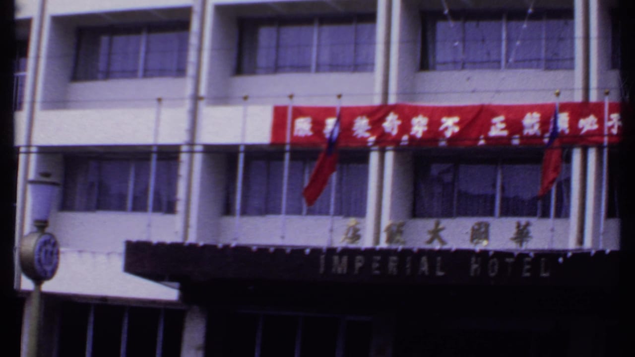 1969 India frame showing a wider Imperial Hotel facade with a red Chinese-character banner, English canopy sign, street clock, windows, and entrance shadows.