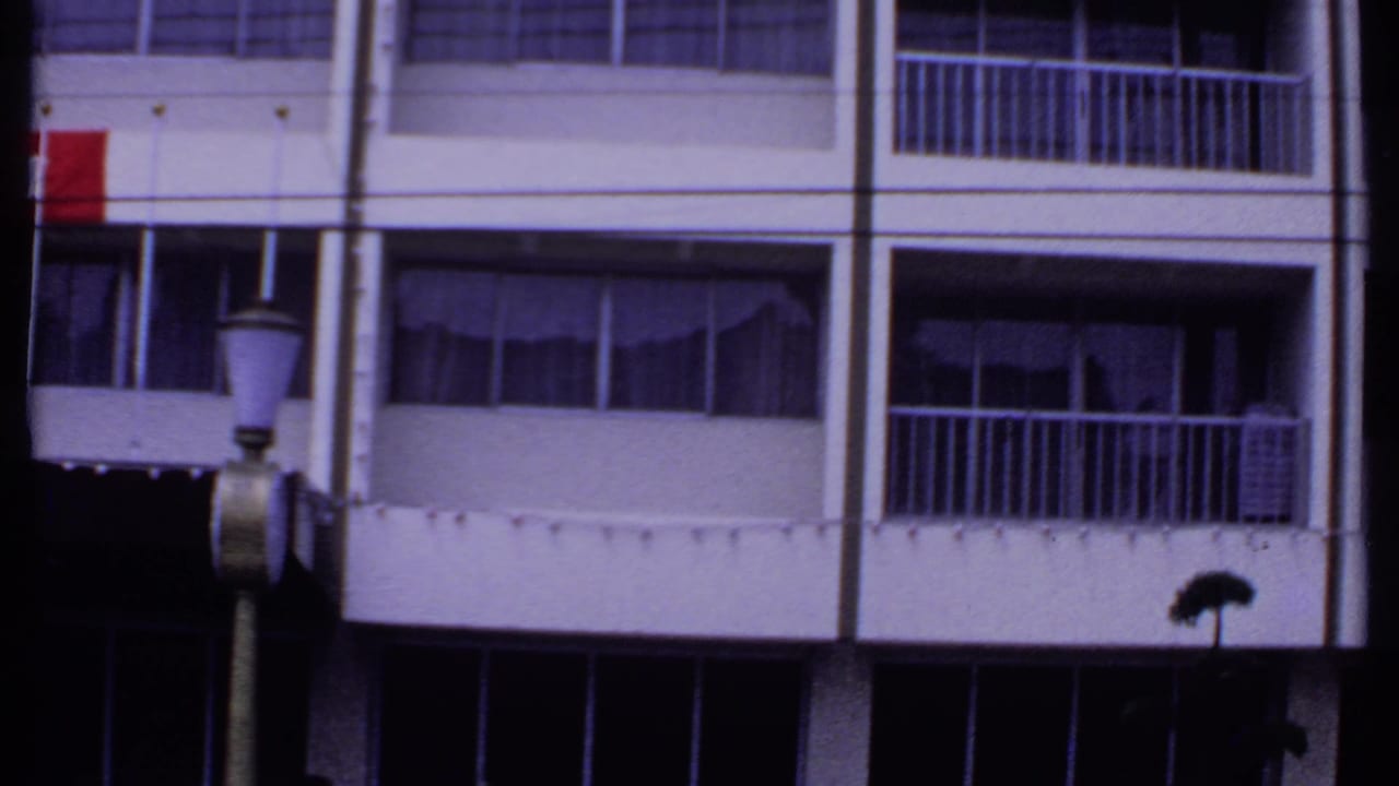 1969 India 8mm frame showing a commercial or residential facade with balconies, curtained windows, foreground lamp, and partial red signage.