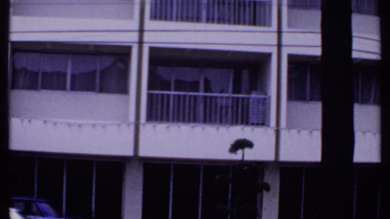 1969 India home-movie frame of a multi-story facade with balcony rails, curtained windows, dark storefront level, and a street lamp silhouette.