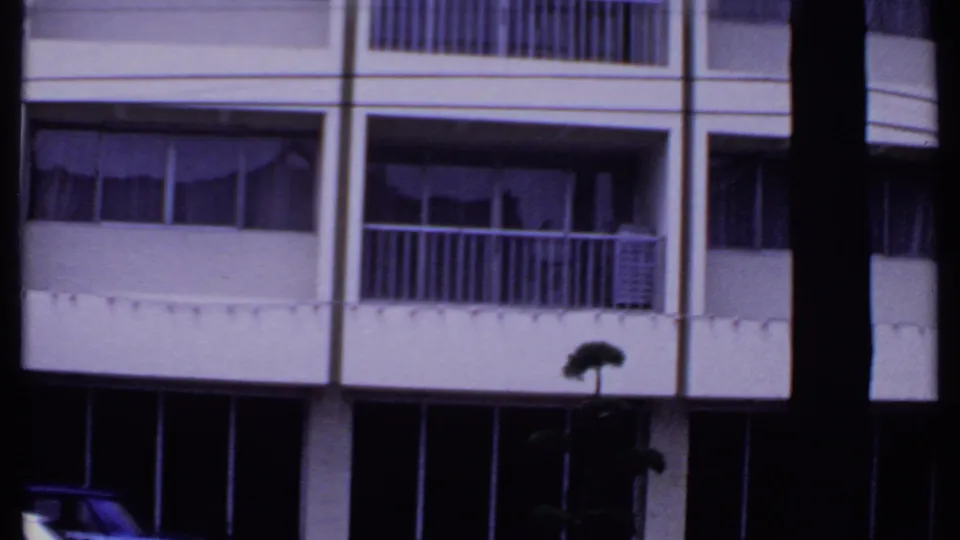 1969 India home-movie frame of a multi-story facade with balcony rails, curtained windows, dark storefront level, and a street lamp silhouette.