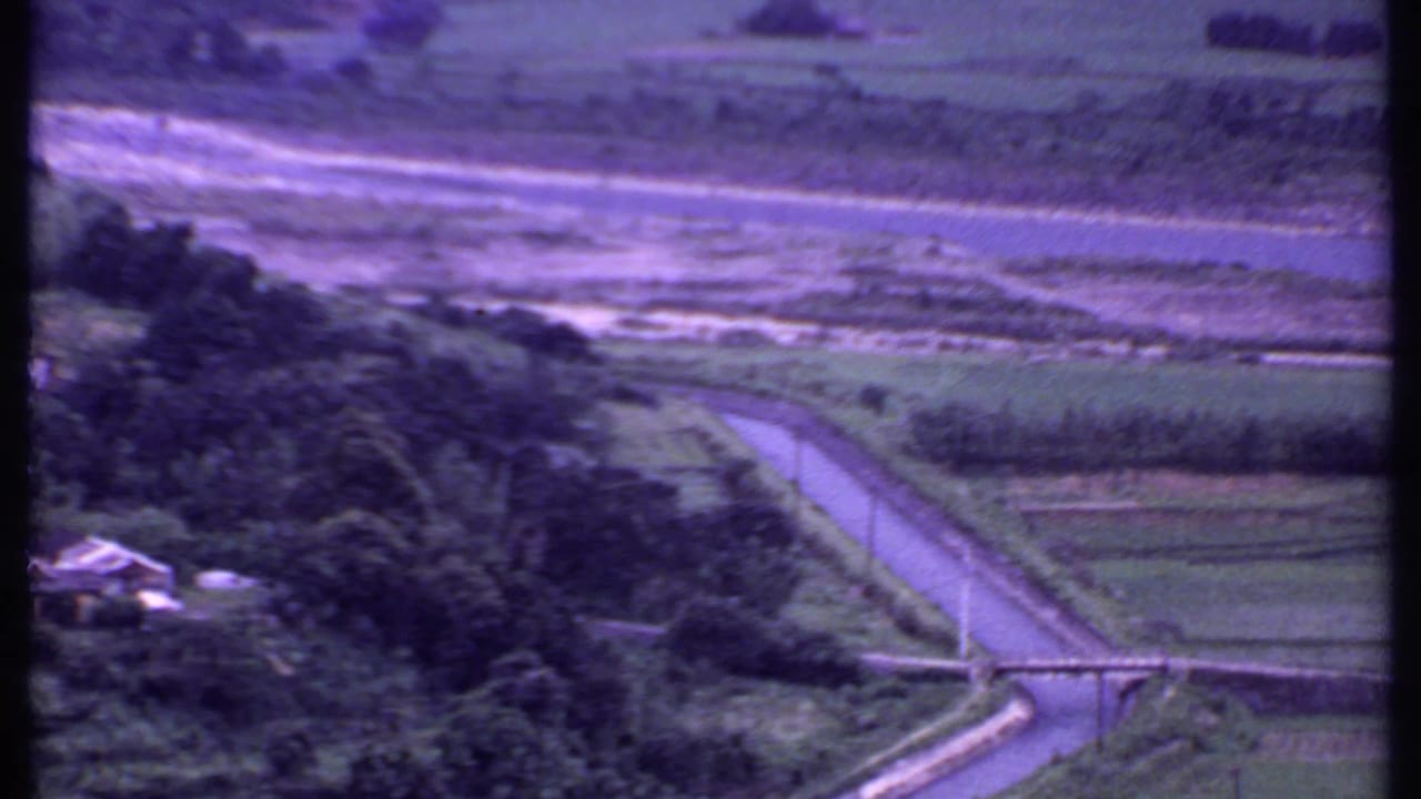 1969 India aerial frame showing a curving road, green fields, trees, a small structure, and water or shoreline beyond.