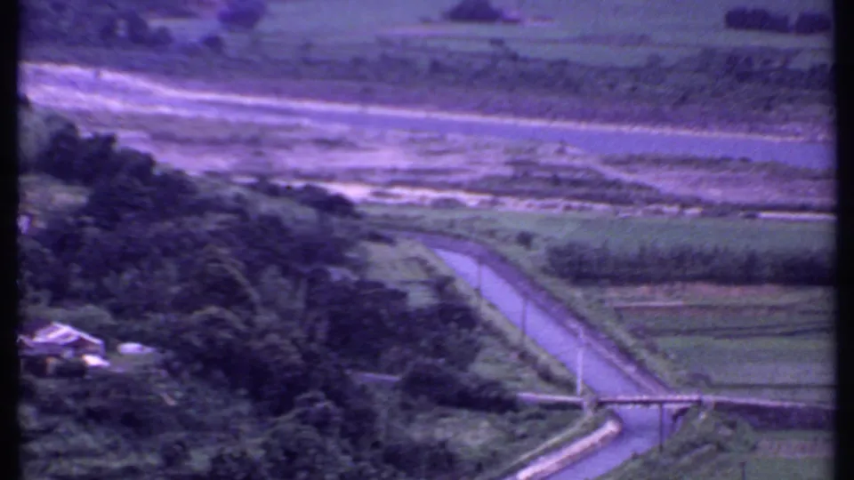 1969 India aerial frame showing a curving road, green fields, trees, a small structure, and water or shoreline beyond.