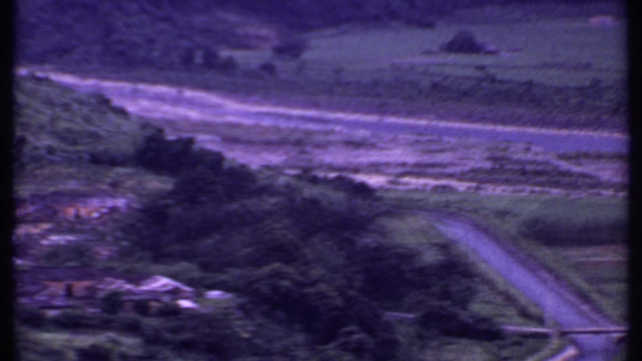 1969 8mm view over a rural Indian hillside village, with a road, small bridge, buildings, trees, and fields under a purple film cast.