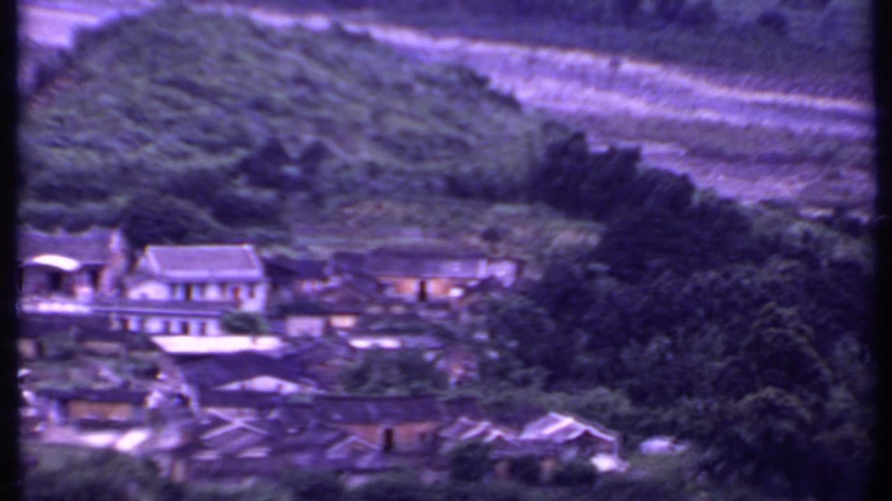 Clustered buildings sit below green hills, with trees and a pale riverbed behind them.