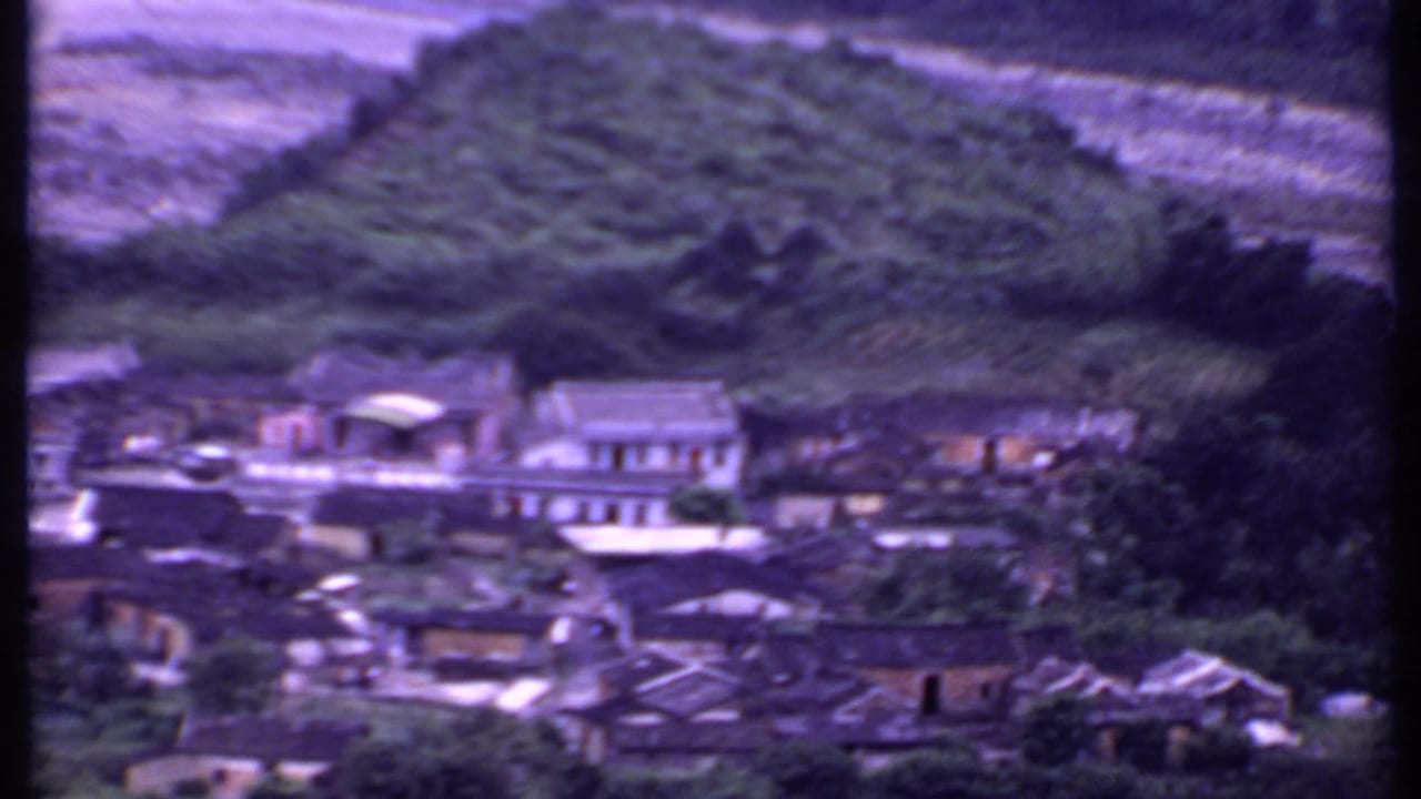 Soft 8mm frame of a rural Indian village in 1969, with tiled roofs, larger buildings, trees, and a cultivated hillside beyond.