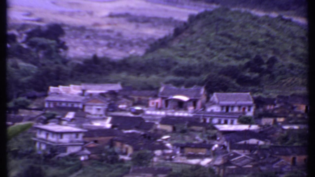 High-angle 1969 8mm view of rural Indian buildings among trees, with a cultivated hill and pale riverbed behind the settlement.
