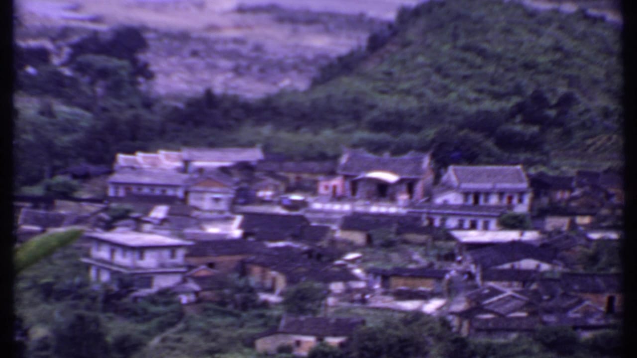 1969 India home-movie frame showing a hillside village among trees and misty mountain slopes, with soft focus and purple color shift.