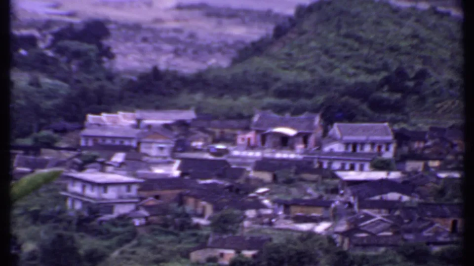 1969 India home-movie frame showing a hillside village among trees and misty mountain slopes, with soft focus and purple color shift.