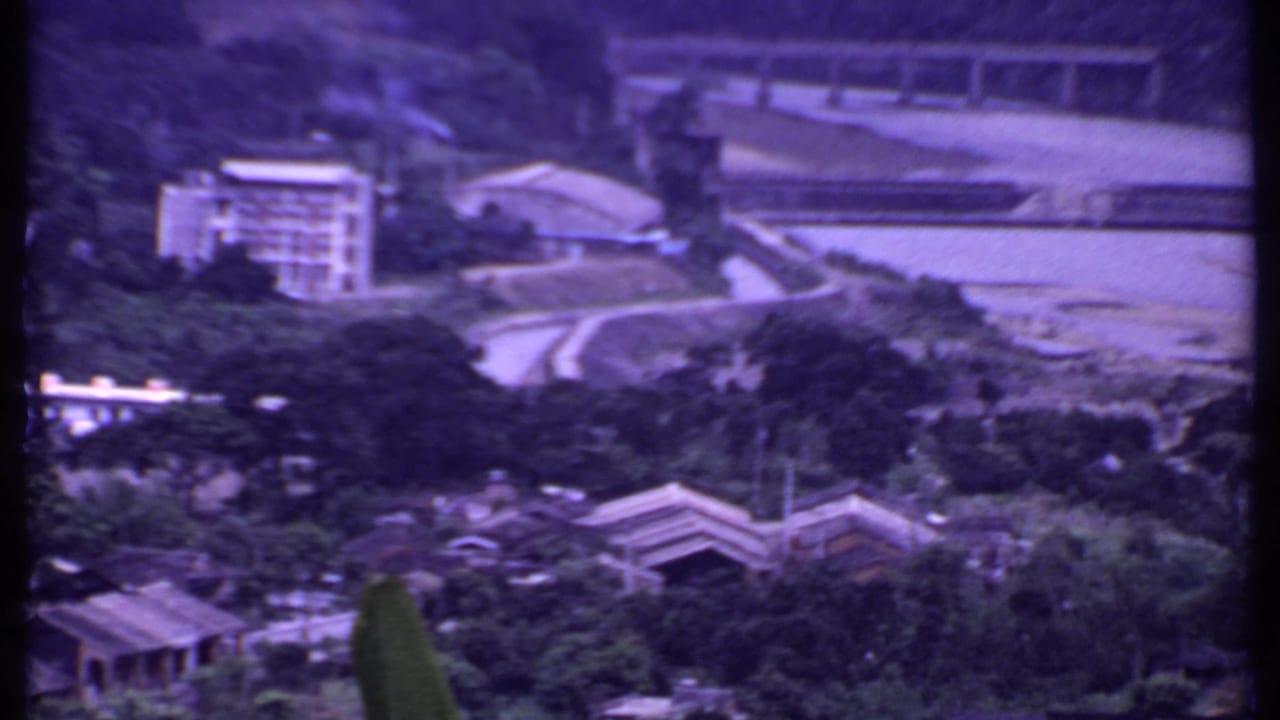 1969 India 8mm frame with a distant building, road, bridge, wooded slope, and water in a purple-tinted home-movie image.