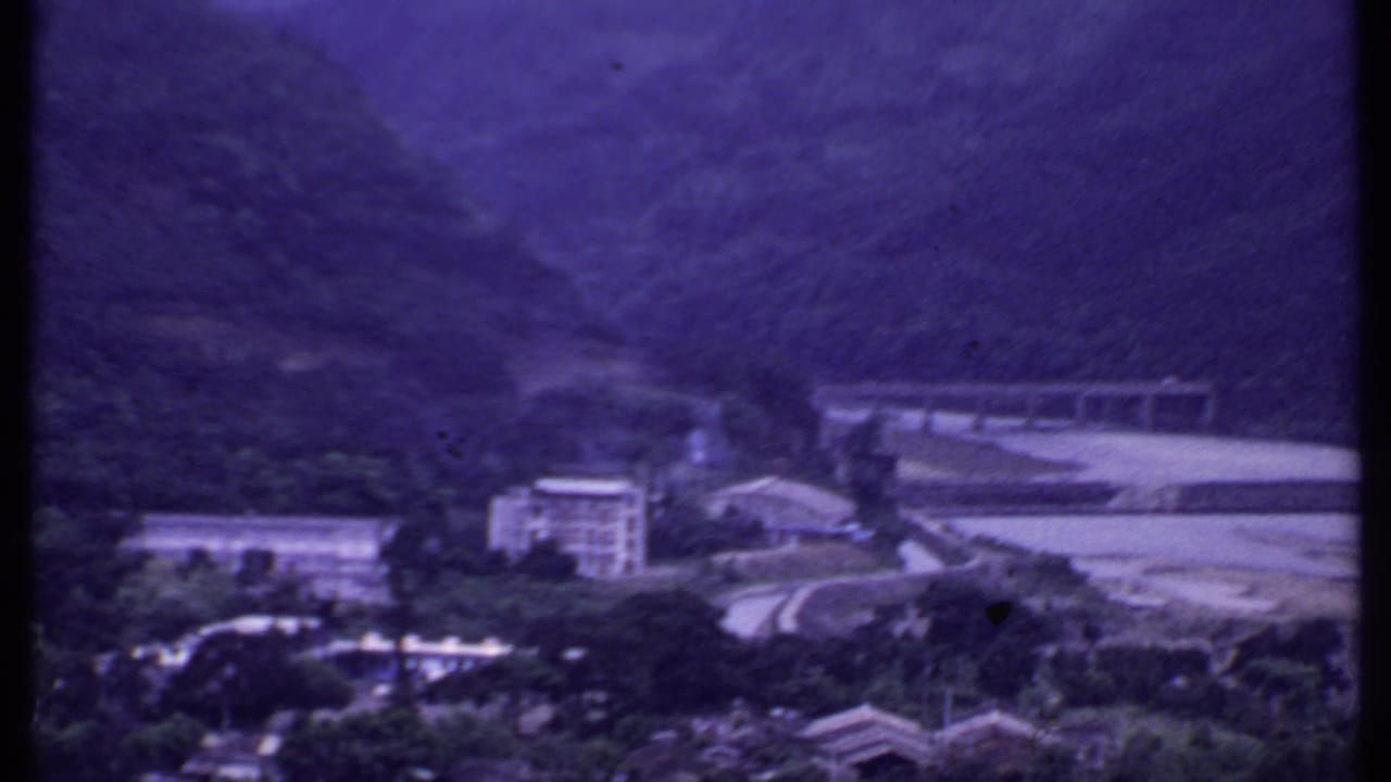 1969 India 8mm frame showing the bridge and road from farther back, with buildings, water, and wooded hills in view.