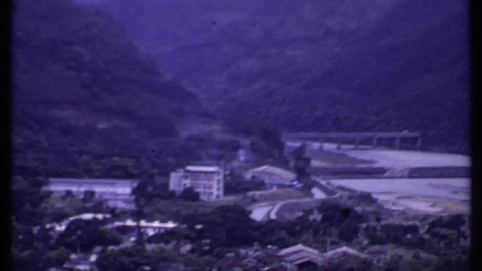 1969 India 8mm frame showing the bridge and road from farther back, with buildings, water, and wooded hills in view.