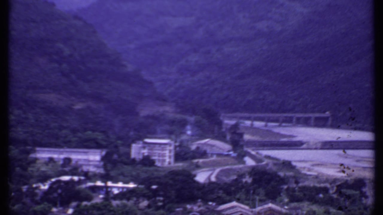 1969 India 8mm view of a mountain valley with a concrete bridge, pale riverbed, hillside vegetation, and riverside buildings.