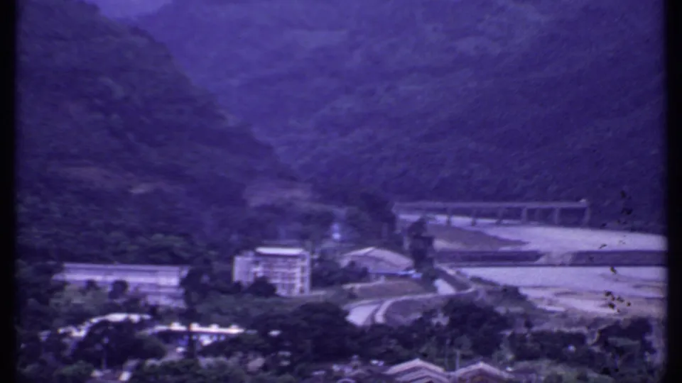 1969 India 8mm view of a mountain valley with a concrete bridge, pale riverbed, hillside vegetation, and riverside buildings.