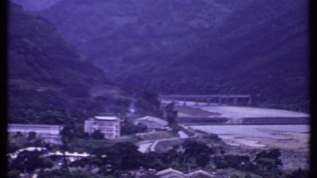 Mountain valley view with long bridge, hillside, riverbank, and buildings.