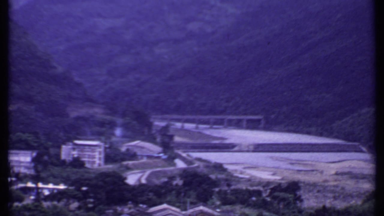 1969 India 8mm frame of a bridge crossing a pale riverbed below steep green hills, with a building at the valley edge.