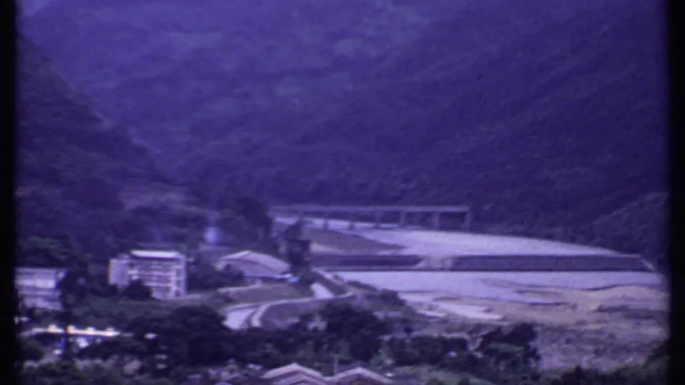 1969 India 8mm frame of a bridge crossing a pale riverbed below steep green hills, with a building at the valley edge.