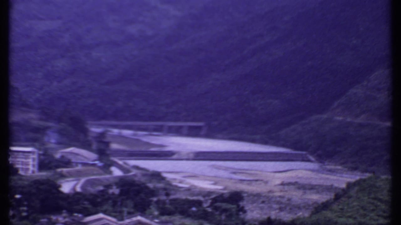 1969 India 8mm frame with the bridge and riverbank framed by steep hills, trees, and a partly visible building at the edge.