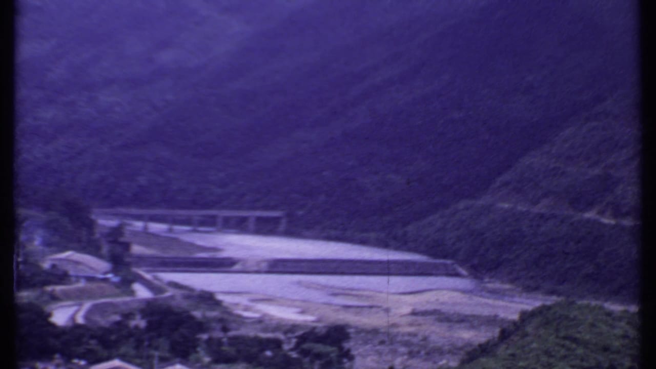 Wide mountain valley with river and pale concrete span in the distance.