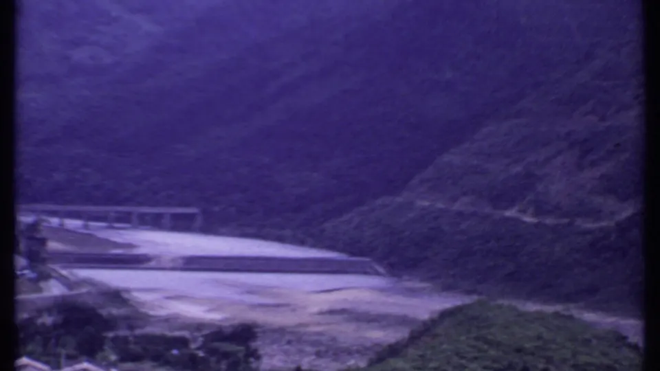 1969 India 8mm view of a mountain river valley with a pale dam or bridge and dark green slopes in the background.