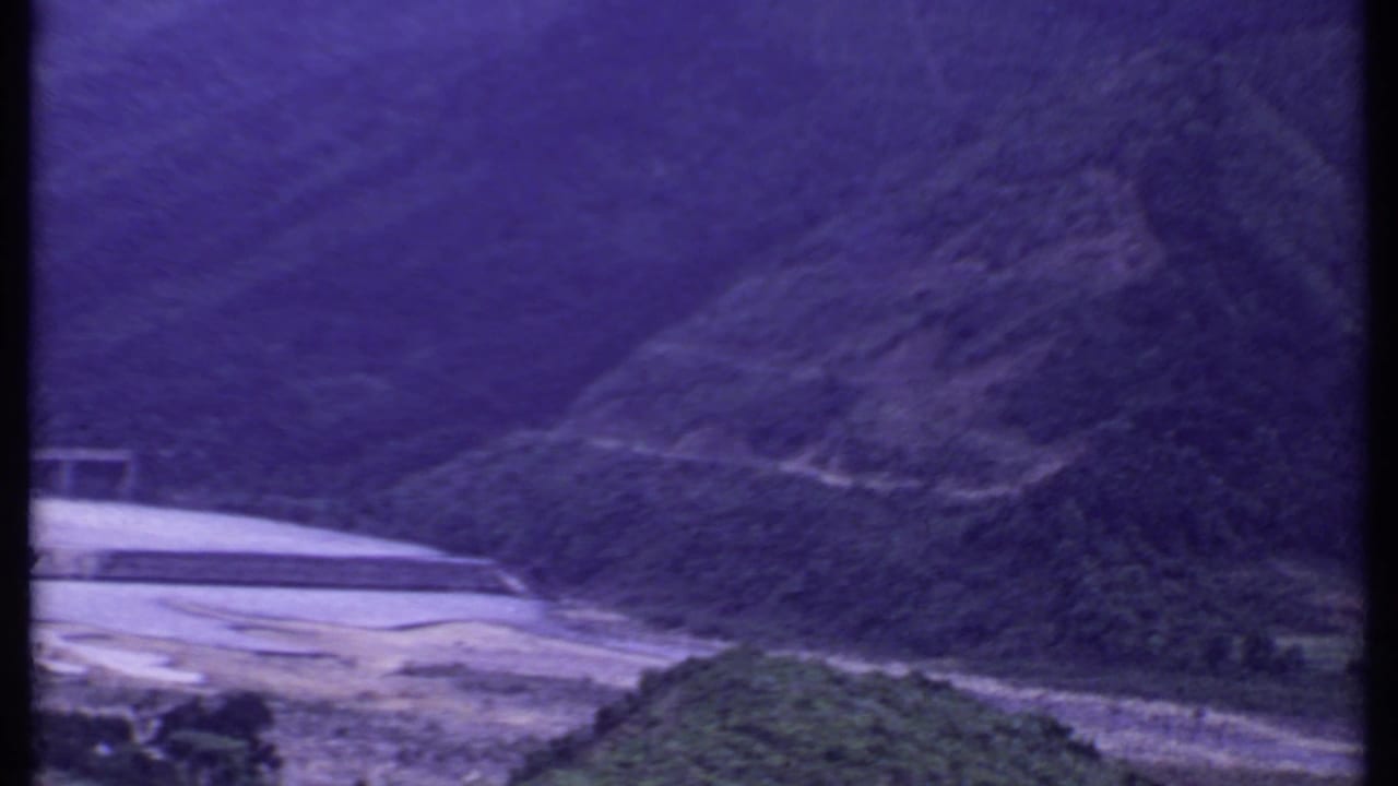 Purple-shifted 8mm frame from India showing a hillside above a riverbank with part of a concrete dam or bridge at left.