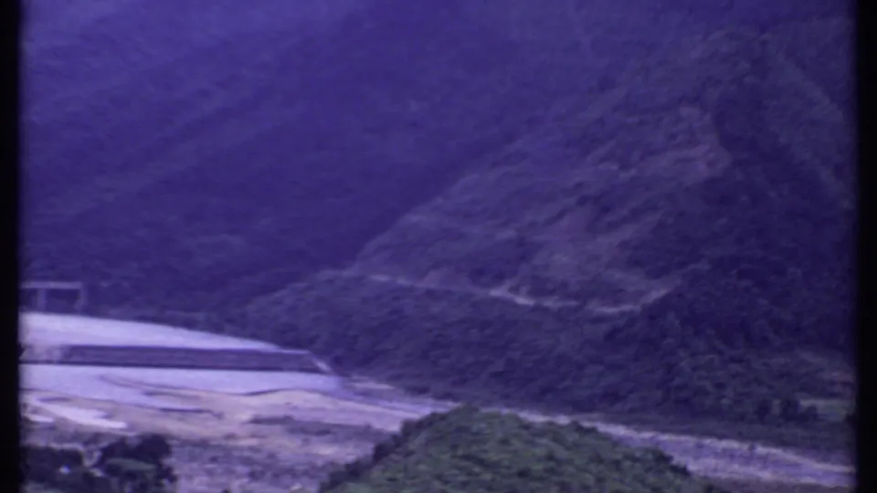 Purple-shifted 8mm frame from India showing a hillside above a riverbank with part of a concrete dam or bridge at left.