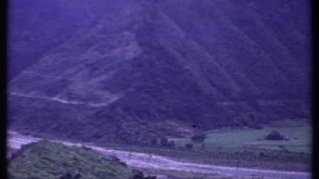 1969 India 8mm frame showing a pale road curving through a green valley below dark, forested hills and a purple color cast.