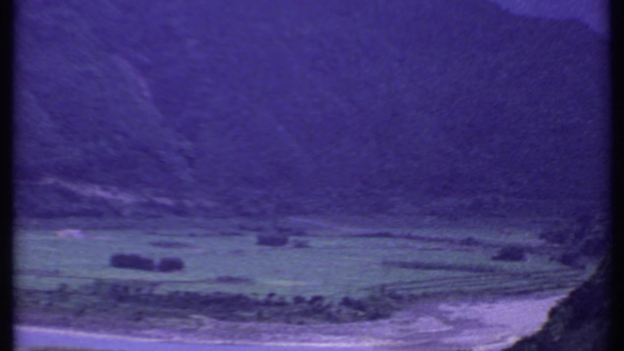 Purple-tinted mountain valley with fields and a pale riverbed below.