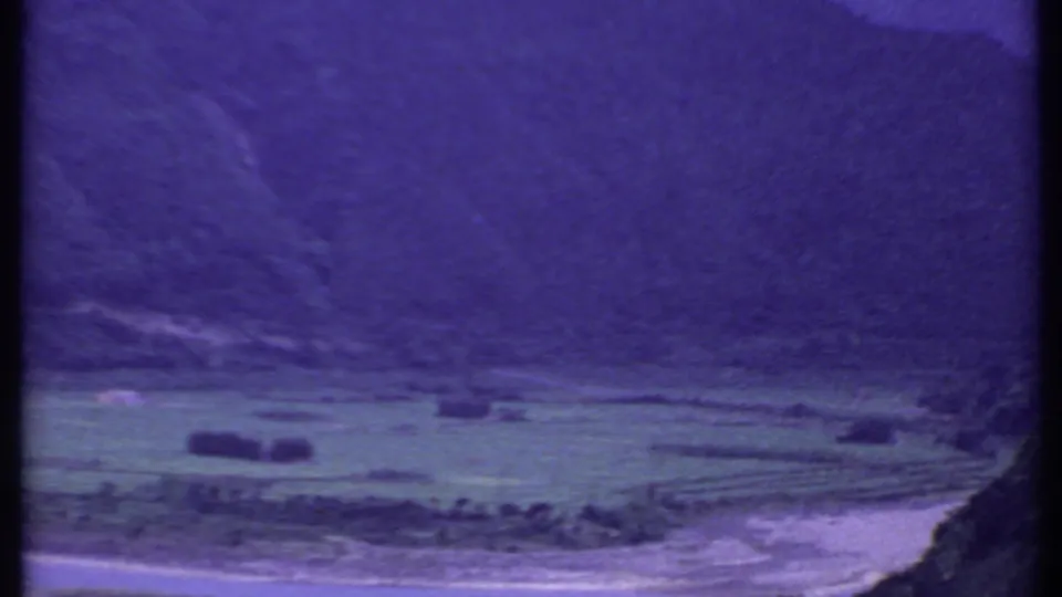 1969 8mm view in India showing a mountain valley, green fields, and a pale riverbed under a purple color shift.