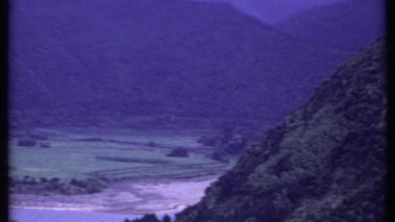 1969 India home-movie frame of a broad valley with terraced fields, a pale river channel, and forested hillside.