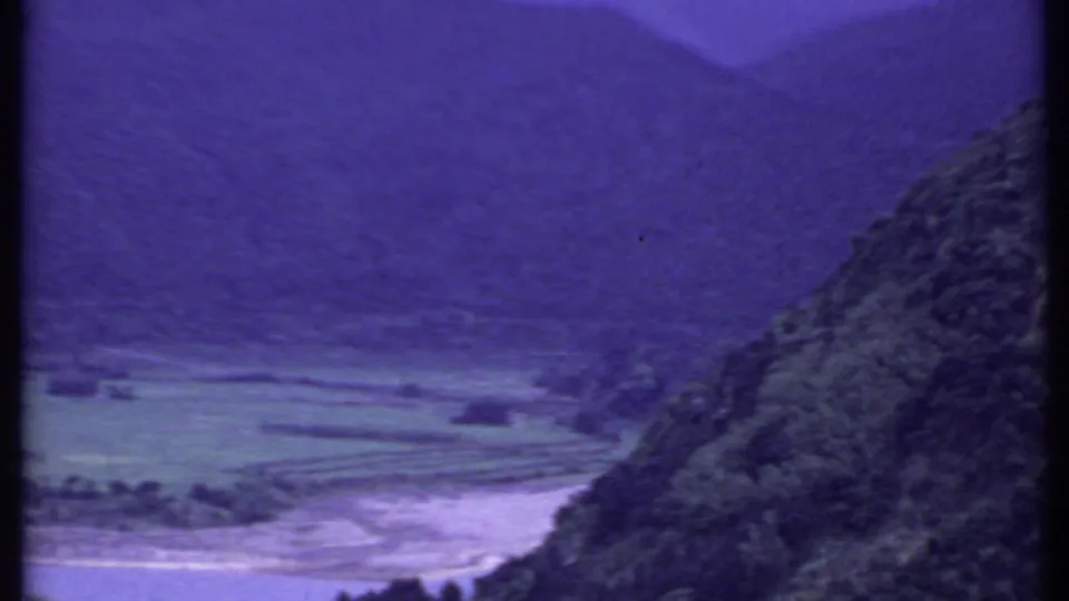 1969 India home-movie frame of a broad valley with terraced fields, a pale river channel, and forested hillside.