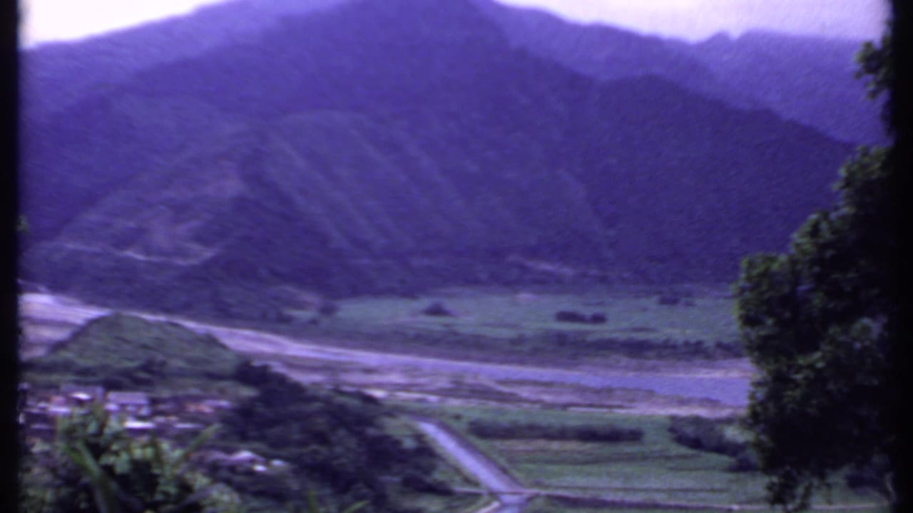 1969 8mm frame from India showing a road entering a valley with small buildings, trees, fields, and steep mountains beyond.