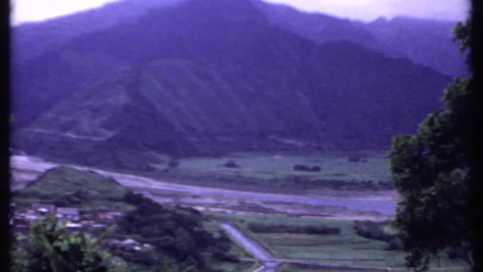1969 8mm frame from India showing a road entering a valley with small buildings, trees, fields, and steep mountains beyond.