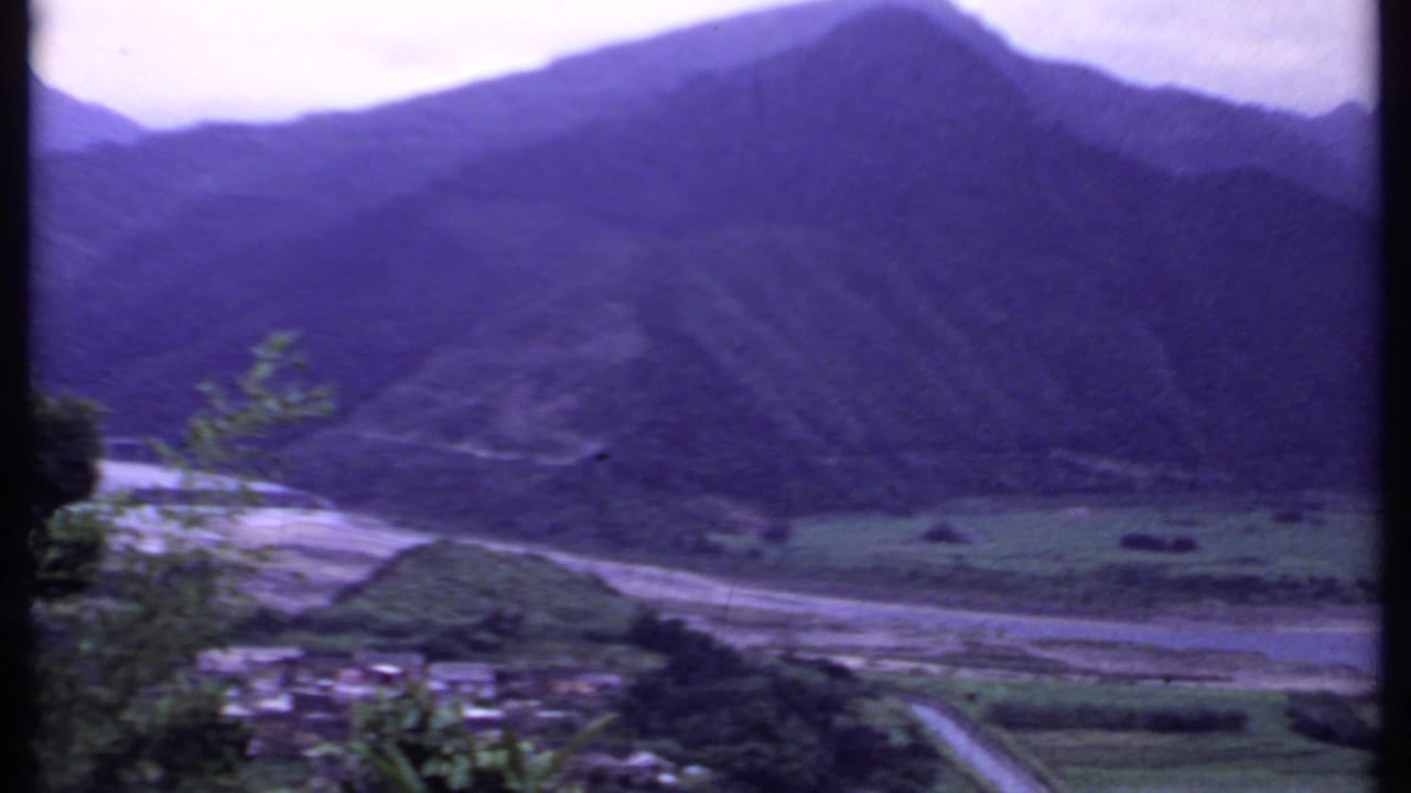 1969 India mountain-valley view with a narrow road, clustered rural buildings, fields, and a pale riverbed beneath dark slopes.