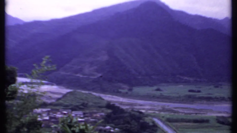 1969 India mountain-valley view with a narrow road, clustered rural buildings, fields, and a pale riverbed beneath dark slopes.