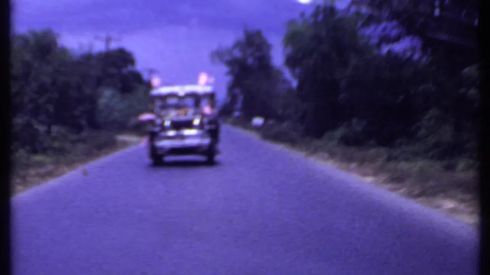 1969 India 8mm frame showing a jeep-style vehicle driving toward the camera on a paved road through wooded hills.