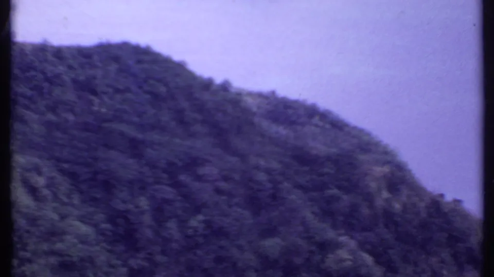 1969 India 8mm frame with a dark green forested mountain slope under a pale, hazy sky.