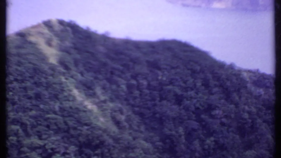 1969 India 8mm frame showing a wooded mountain slope with a lighter clearing or path descending through the vegetation.