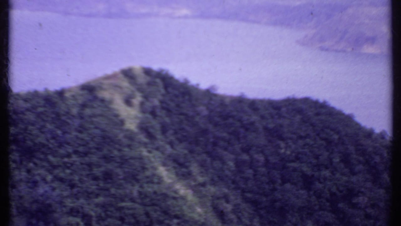 1969 India 8mm frame showing a forested hill in front of a distant body of water under hazy light.