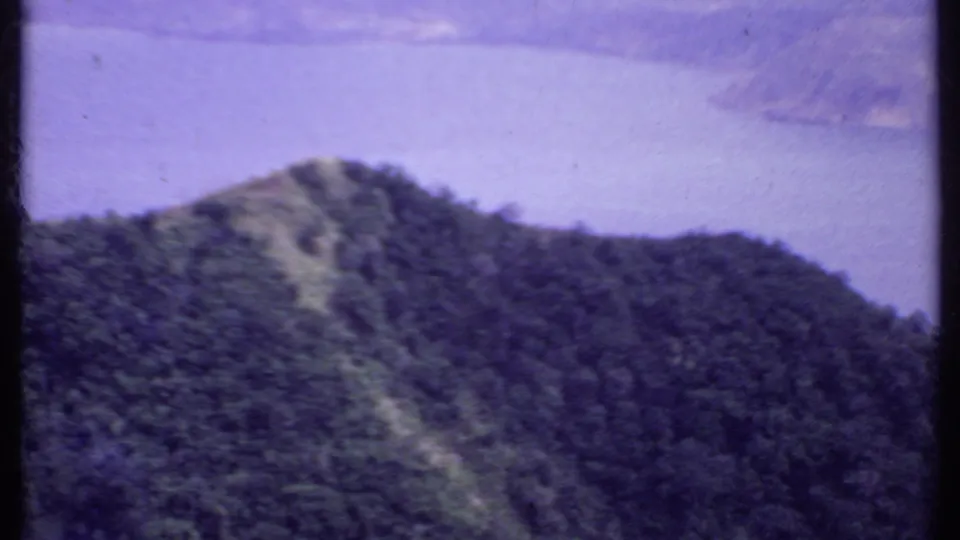 1969 India 8mm frame showing a forested hill in front of a distant body of water under hazy light.