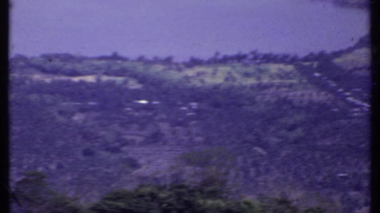 1969 8mm India view over a rural lakeside town or village, with terraced fields, scattered buildings, roads, and hills in the distance.