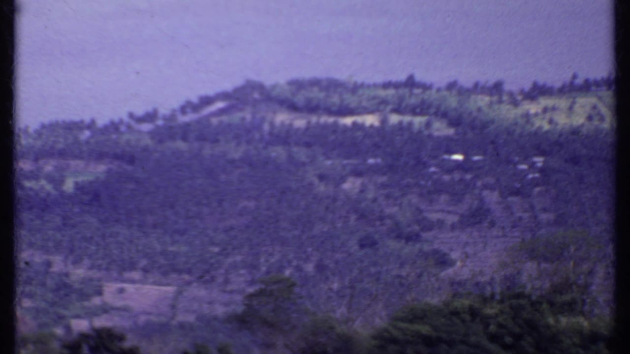 1969 8mm India frame of a distant hillside settlement with cultivated slopes, small structures, vegetation, and a soft hazy mountain backdrop.