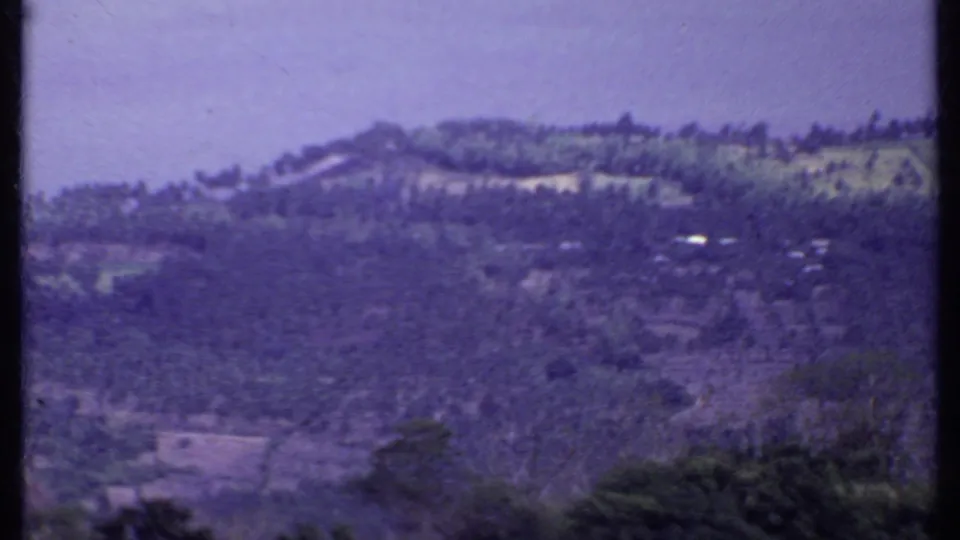 1969 8mm India frame of a distant hillside settlement with cultivated slopes, small structures, vegetation, and a soft hazy mountain backdrop.