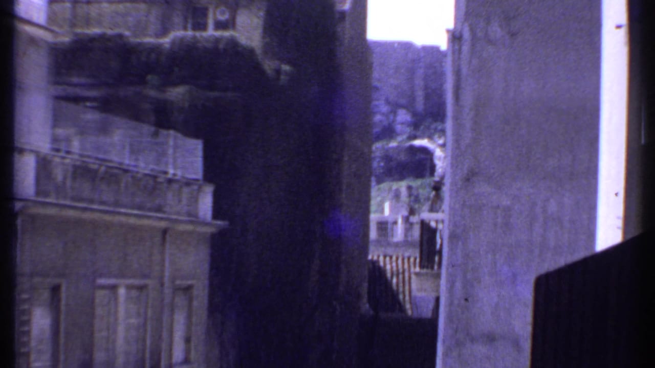 1969 Kenya frame showing a weathered pale facade with balcony rails, windows, tree cover, and a tight passage between buildings.