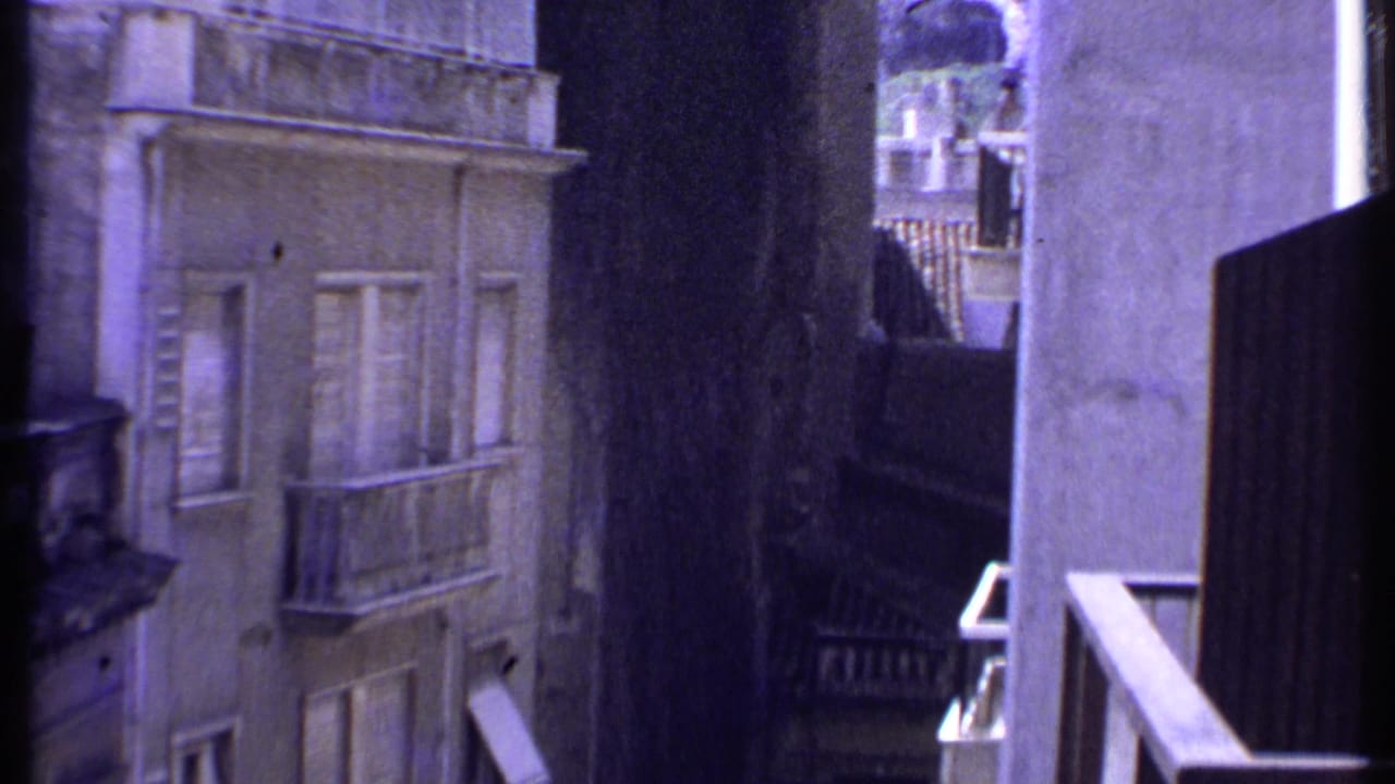 A 1969 Kenya 8mm frame looking through a window toward a narrow alley, pale building facade, balcony, railing, and shadowed foreground edge.